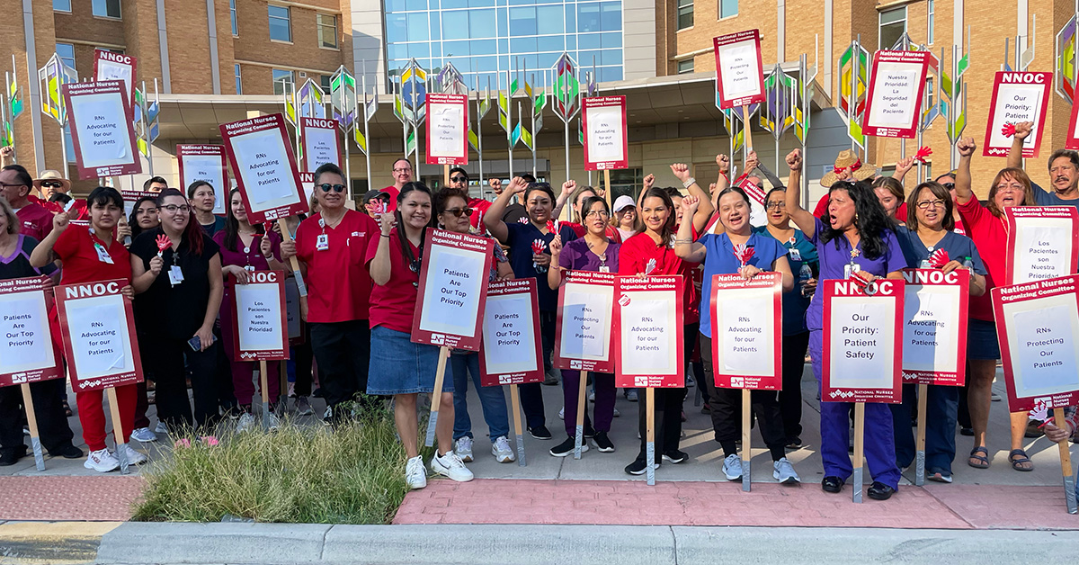 Large group of nurses outside hospital with fists raised, signs that read "Patients are our top priority"