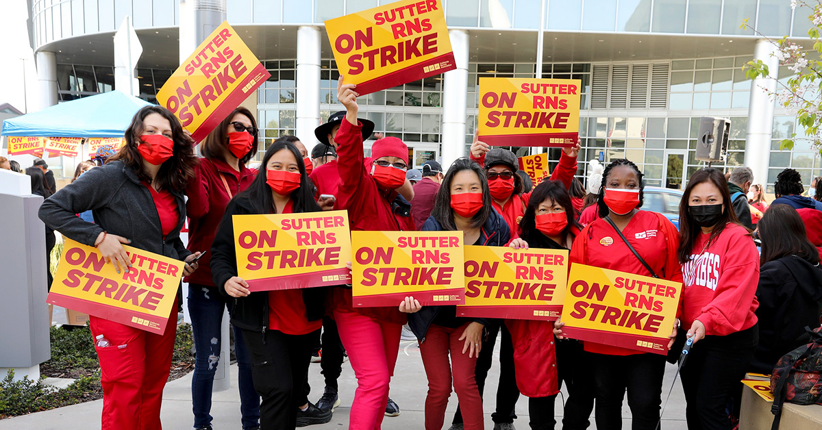 Large group of nurses outside hospital hold signs "On Strike"