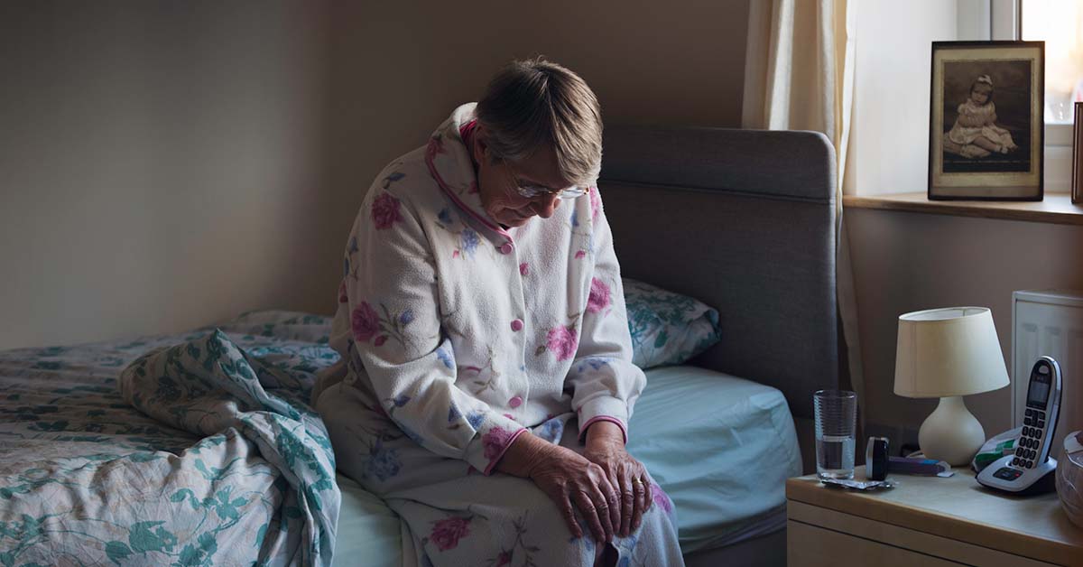 Older woman sits on side of bed with head down