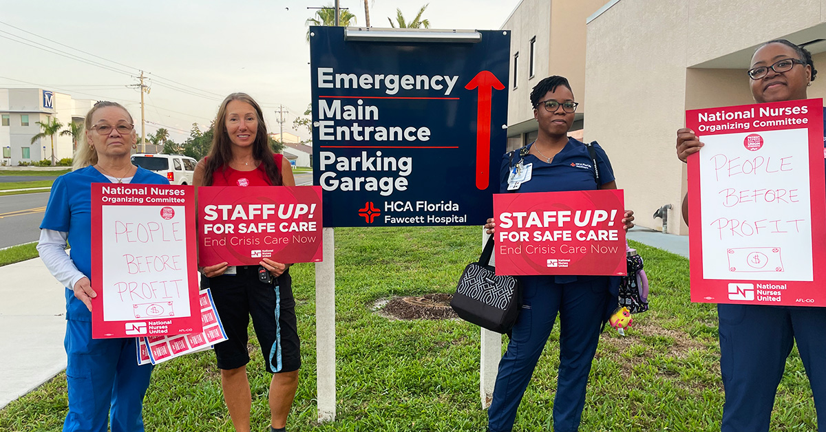 Group of four nurses outside hospital hold signs "People Before Profit" and "Staff Up for Safe Patient Care"