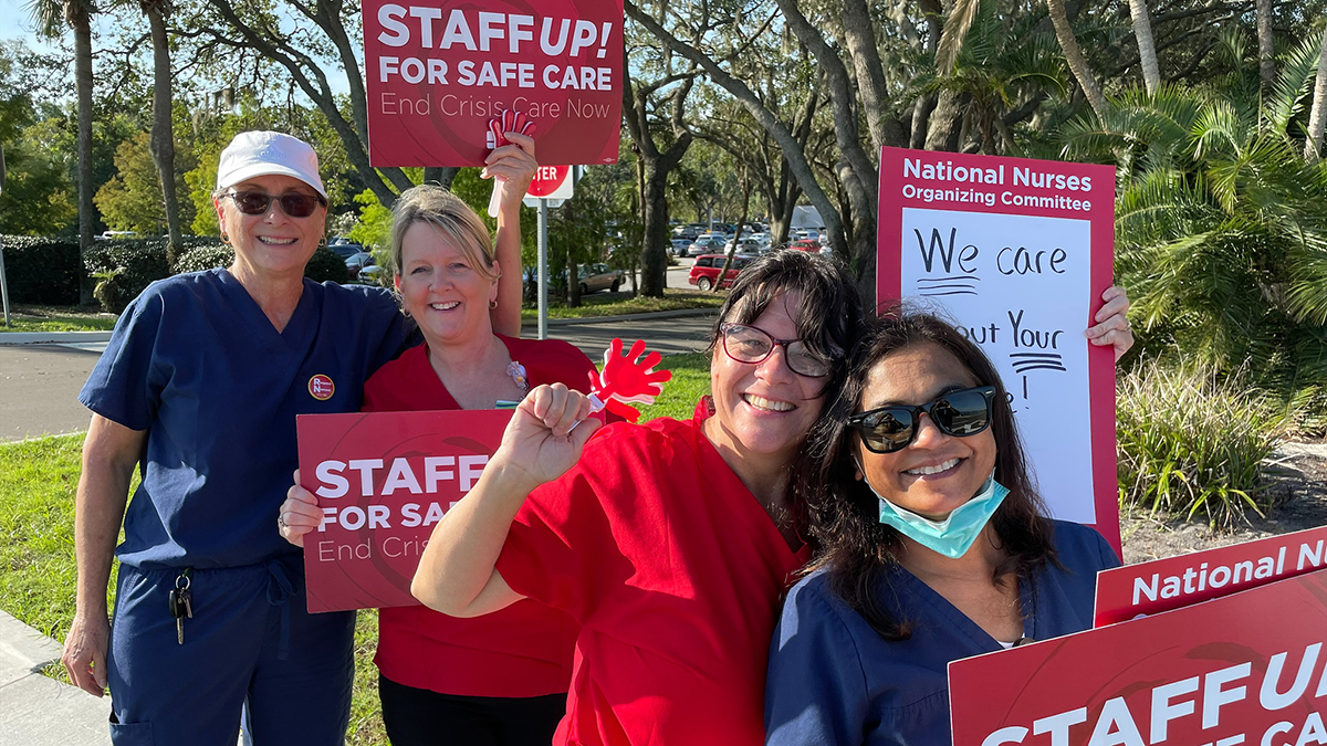 Four nurses outside hold signs "Staff Up for Safe Patient Care"