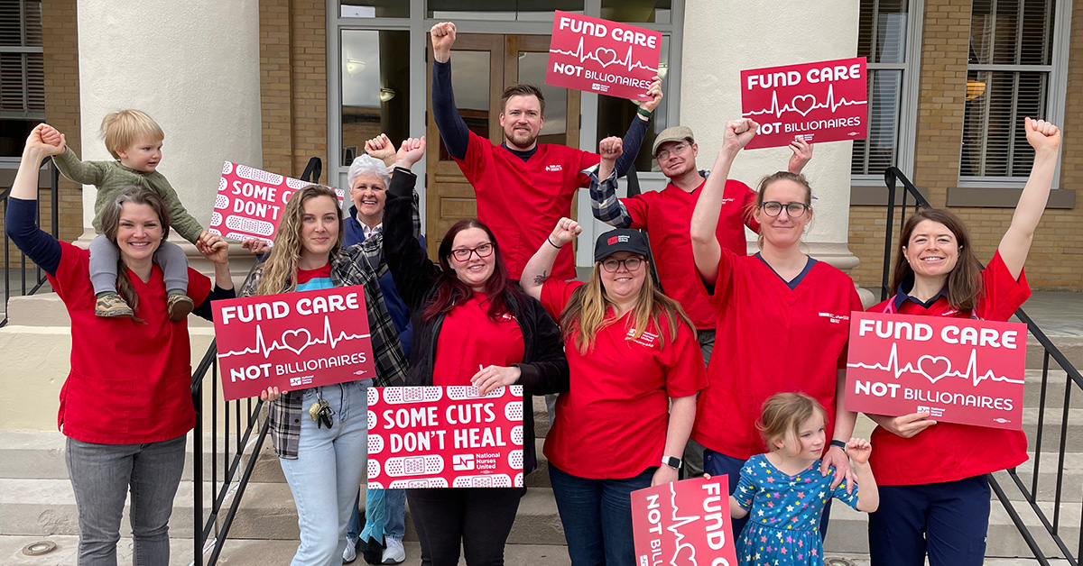 Group of nurses outside in rain clothes holding signs "Some Cuts Don't Heal" and "Fund Care Not Billionaires"