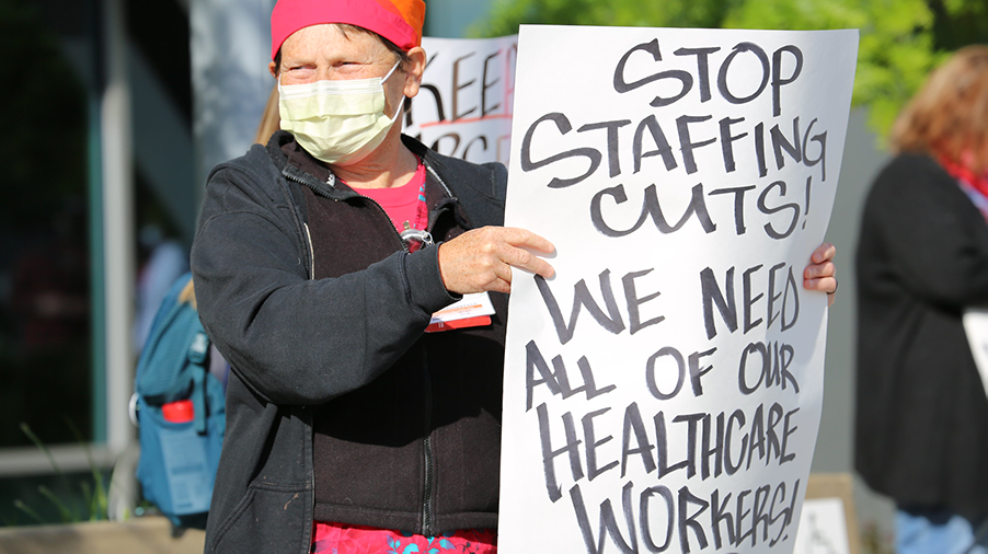 Nurse holds sign "Stop Staffing Cuts! We Need All Health Care Workers"