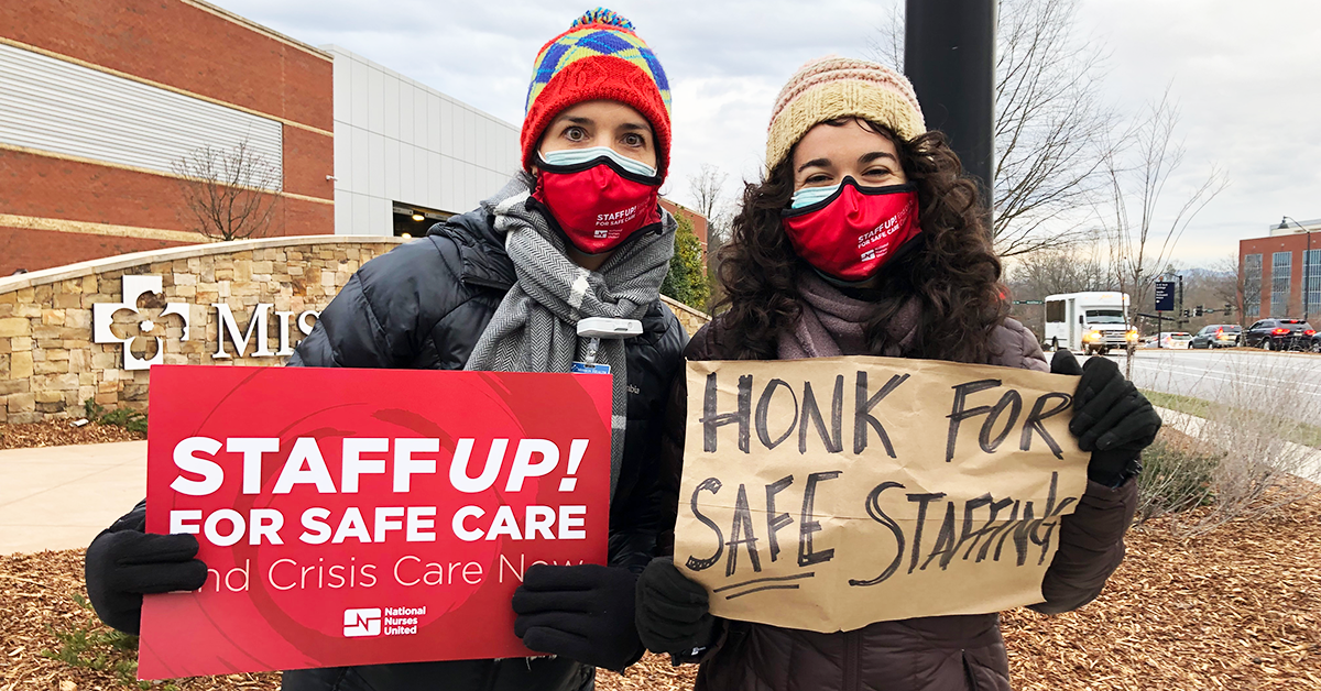 Two nurses outside MIssion Hospital holding signs: "Staff up! for safe care. End crisis care now." and "Honk for safe staffing."