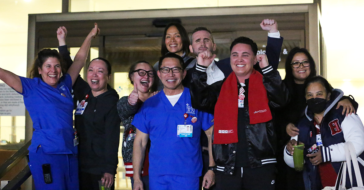 Large group of nurses outside hospital, smiling, with raised fists