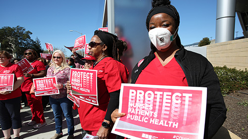Masked nurses holds sign "Protect Nurses, Protect Patients, Protect Public Health from Covid-19"