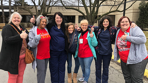 Group of nurses outside, arms around each other, giving thumbs up