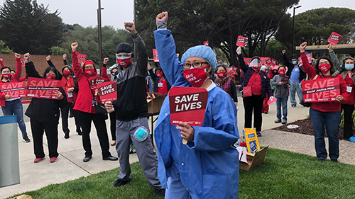 Large group of nurses outside with raised fists, holding various signs that start with "Save Lives"