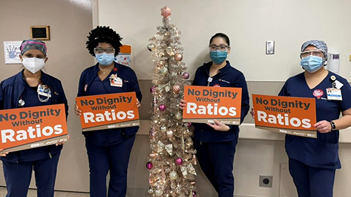 Four nurses inside hospital hold signs "No Dignity, No Ratios"