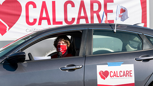 Person in car in front of large CalCare banner, speaking into microphone at car rally.