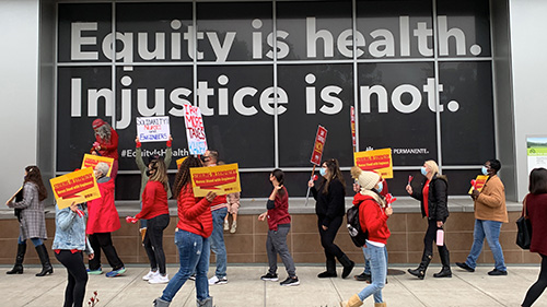 Nurses picketing outside building. Text "Equity is Health, Injustice is Not" displays on side of building.