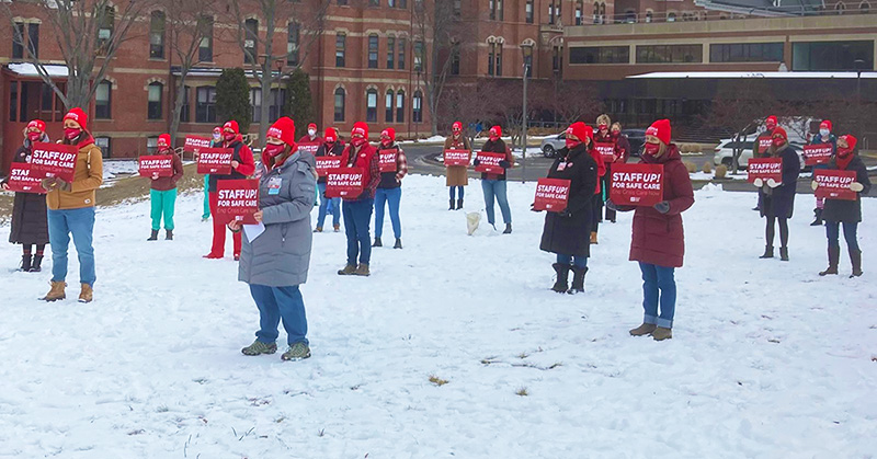 Large group of nurses outside in snow hold signs "Staff Up for Safe Patient Care"