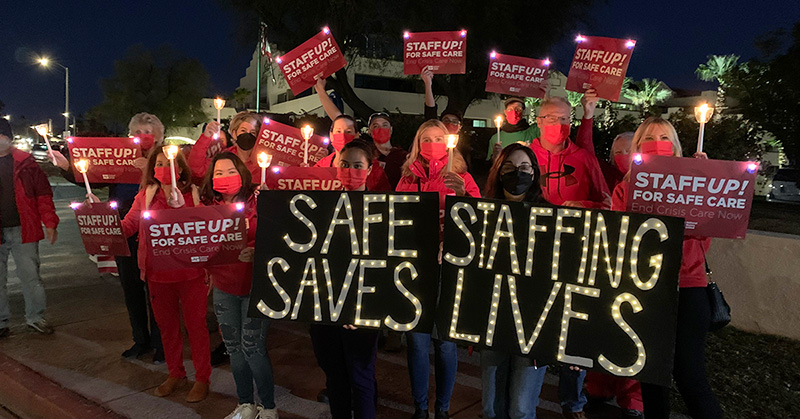 Large group of nurses holding candlelight vigil, holding signs calling for safe staffin