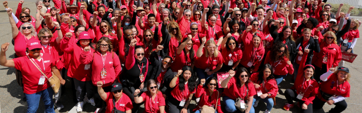 Large group of nurses outside, wearing red, with raised fists