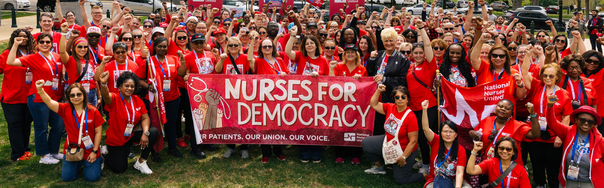 Large group of nurses smiling, with raised fists, wearing red, holding banner "Nurses for Democracy"