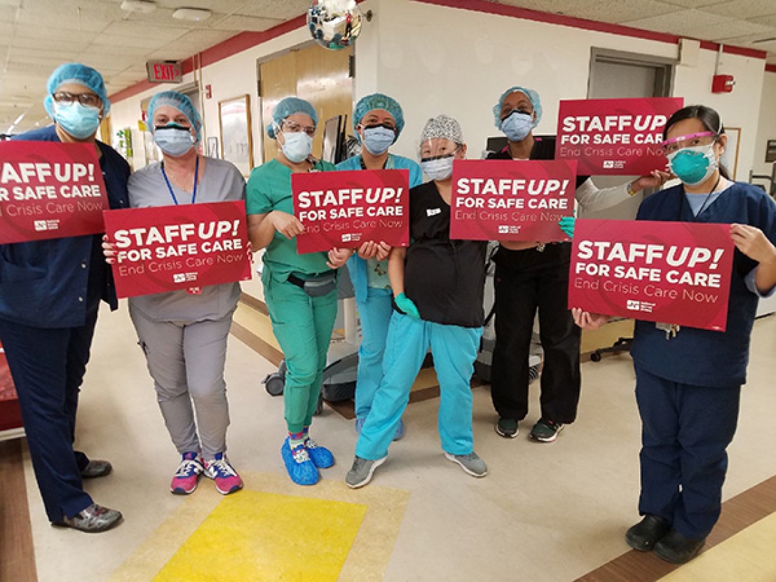 Nurses inside hospital hold signs "Staff Up for Safe Care"