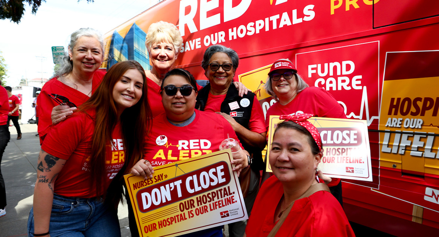 Group of nurses in front of Red Alert tour bus, smiling, holding sign "Don't close our hospital"