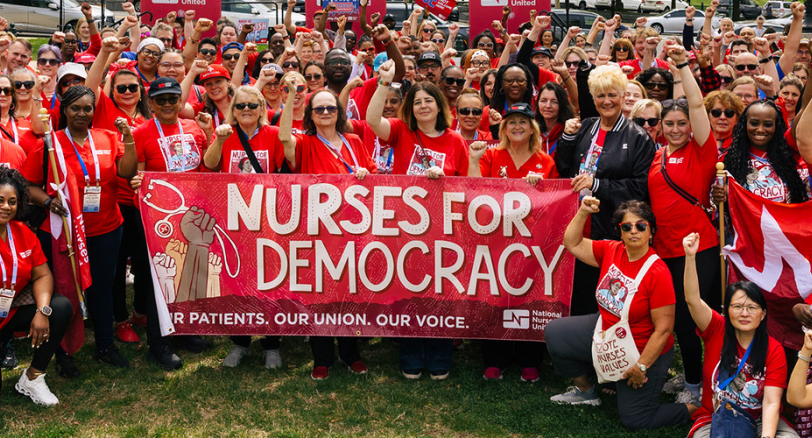 Large group of nurses smiling, with raised fists, wearing red, holding banner "Nurses for Democracy"