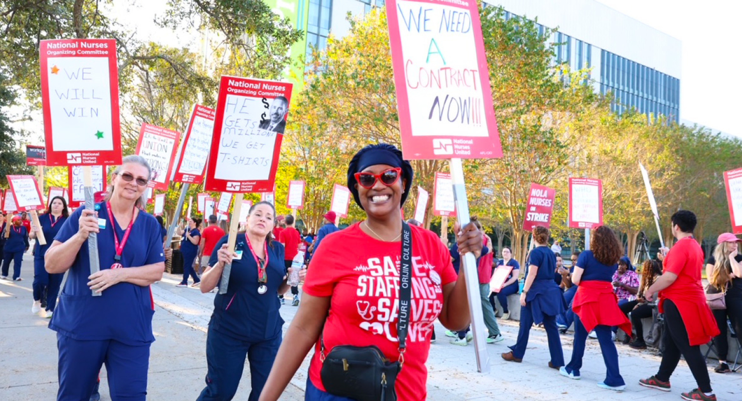 Nurses on picket line, smiling, one holds sign "We Need a Contract NOW!"
