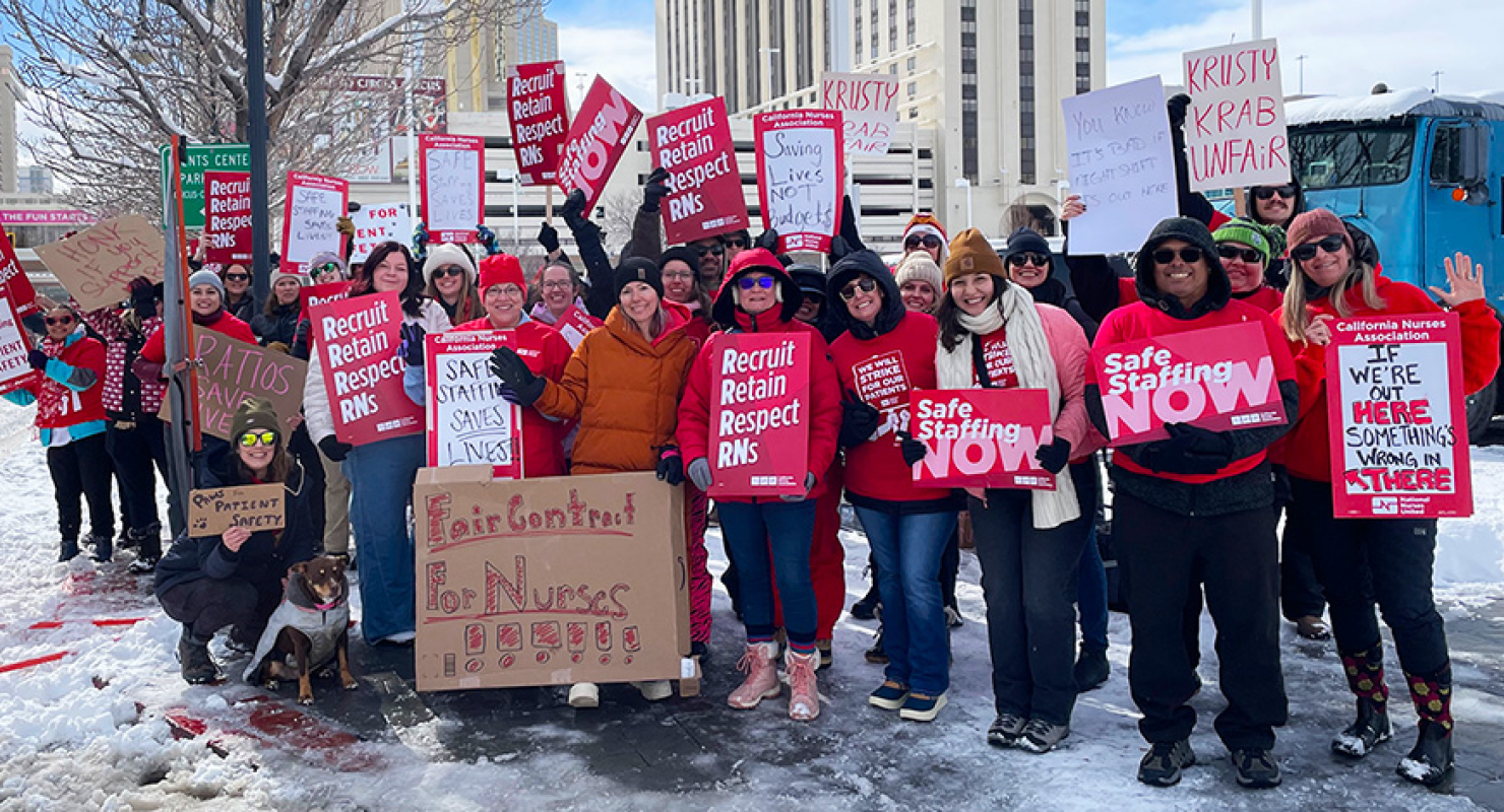 Large group of nurses outside hospital, in cold weather clothes, holding signs including "Safe Staffing NOW" and "Recruit, Retain, Respect RNs"