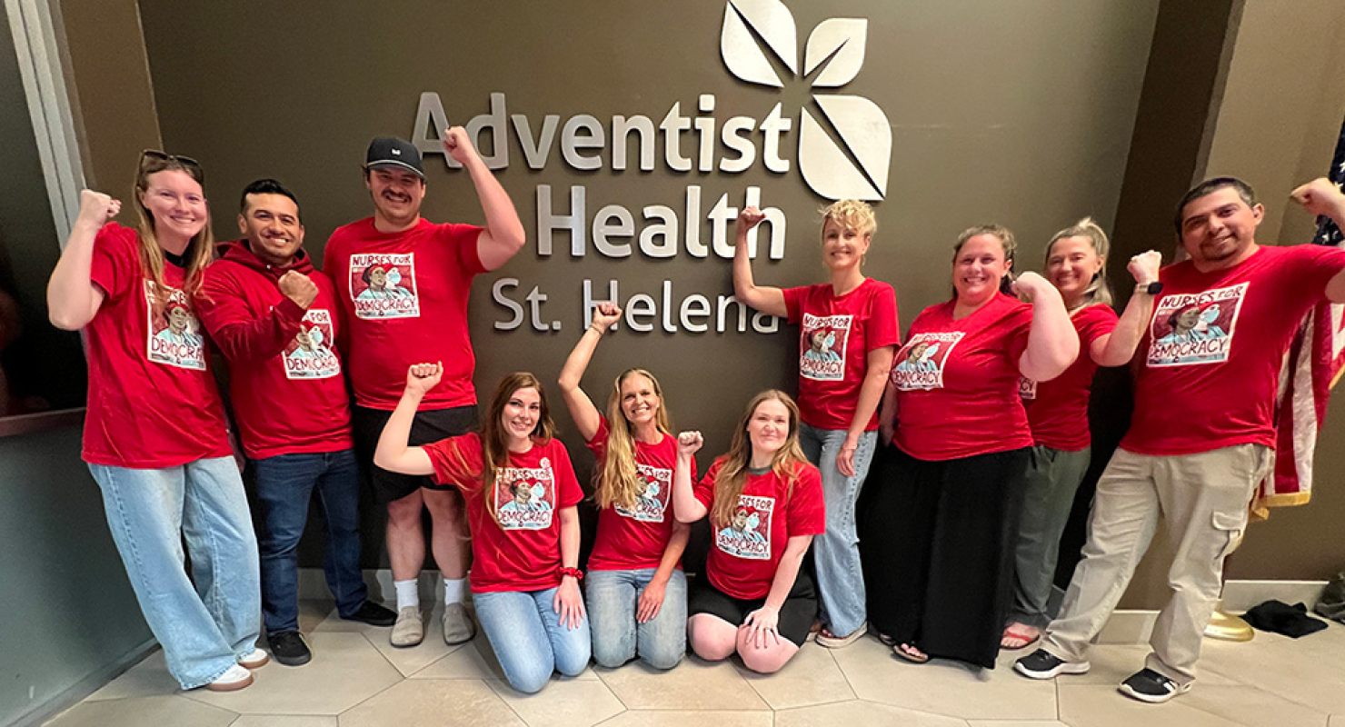 Group of nurses inside, in front of sign "Adventist Health", smiling, wearing red, and with raised fists