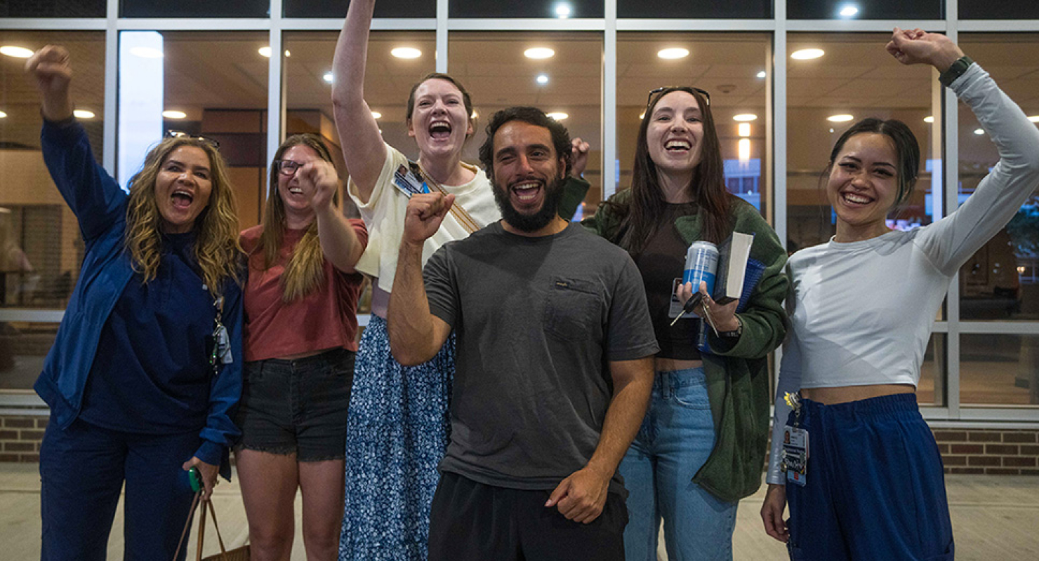 Group of six nurses outside hospital, smiling, with raised fists
