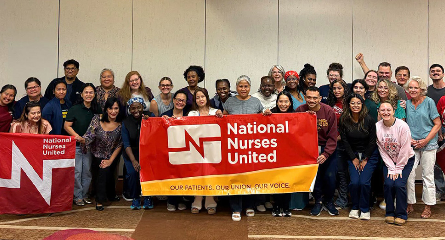 Large group of nurses inside conference room holding banner "National Nurses United"
