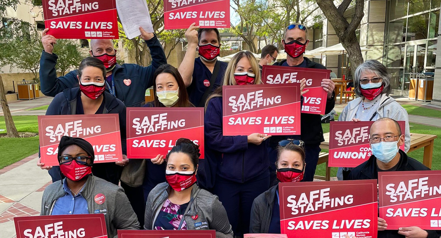Large group of nurses outside hospital hold signs "Staff Up for Safe Care"