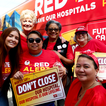 Group of nurses in front of Red Alert tour bus, smiling, holding sign "Don't close our hospital"