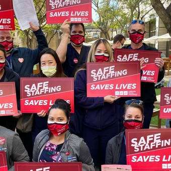 Large group of nurses outside hospital hold signs "Staff Up for Safe Care"