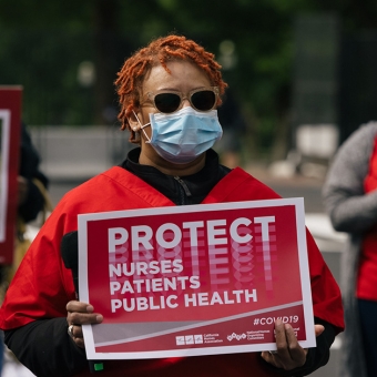 Masked nurse outside holds signs "Protect Nurses, Patients, Public Health"