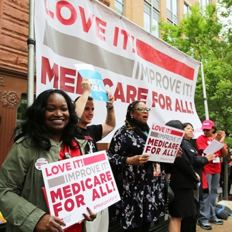 Group of people outside holding signs and standing under banner "Love it! Improve it! Medicare for all!"