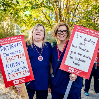 Two nurses smiling and holding signs "Patients Deserve Better, Nurses Deserve Better"