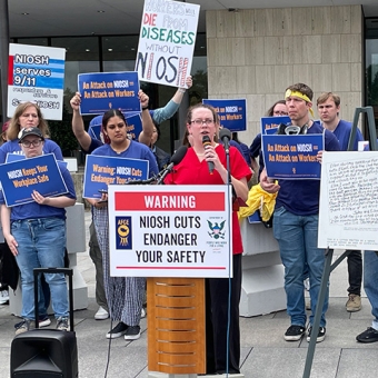 Nurse at podium, behind them health care workers holding signs "An Attack on NIOSH = Attack on workers"
