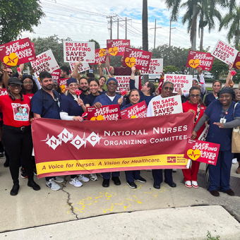Large group of nurses outside, holding banner "National Nurses Organizing Committee" and signs "Safe Staffing Saves Lives"