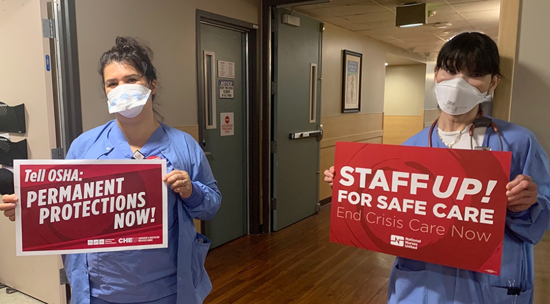 Two nurses inside hospital hold signs calling for permanent ETS and safe staffing