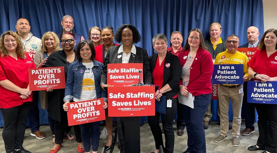 Group of nurses stand at podium holding signs "Safe Staffing Saves Lives"