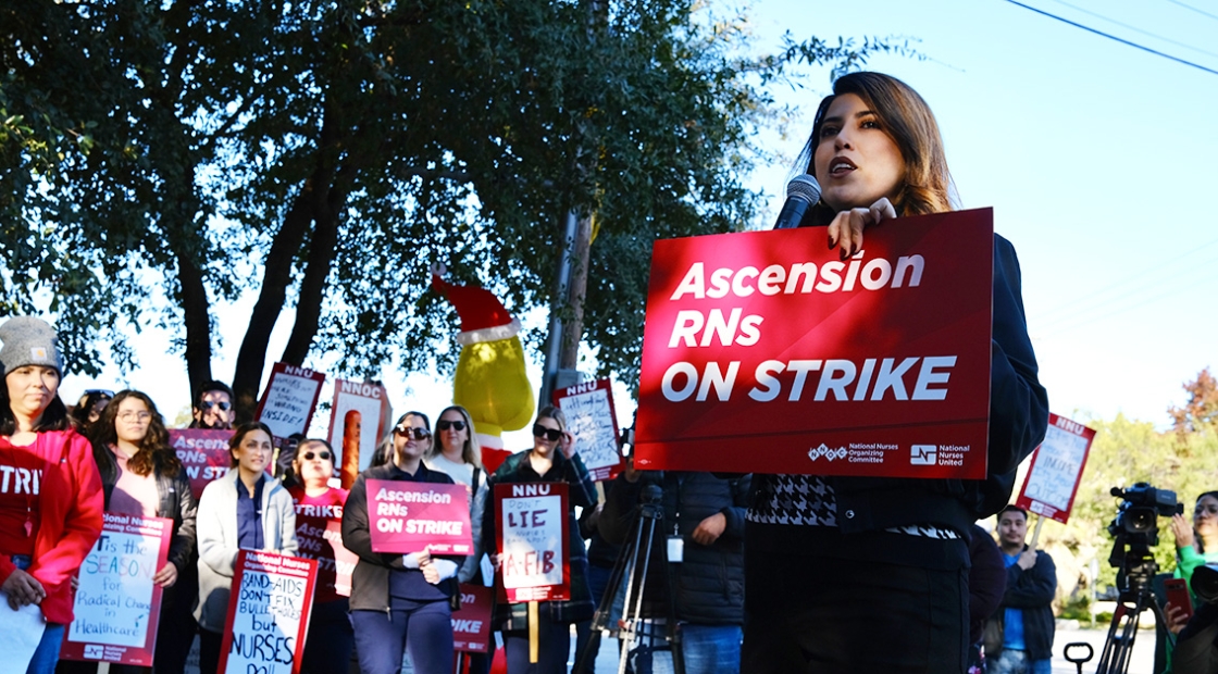 Nurse speaking into microphone holding sign "Ascension RNs On Strike". Other nurses with signs behind.