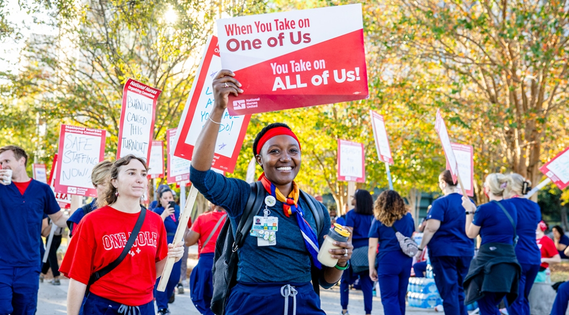 Nurse on picket line holding sign "When You Take On One Of Use, You Take On All Of Us"