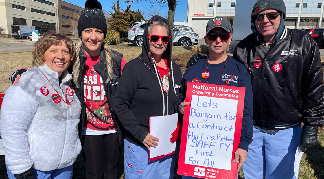 Group of nurses outside hospital, one holding sign "Let's bargain for a contract that puts safety first"