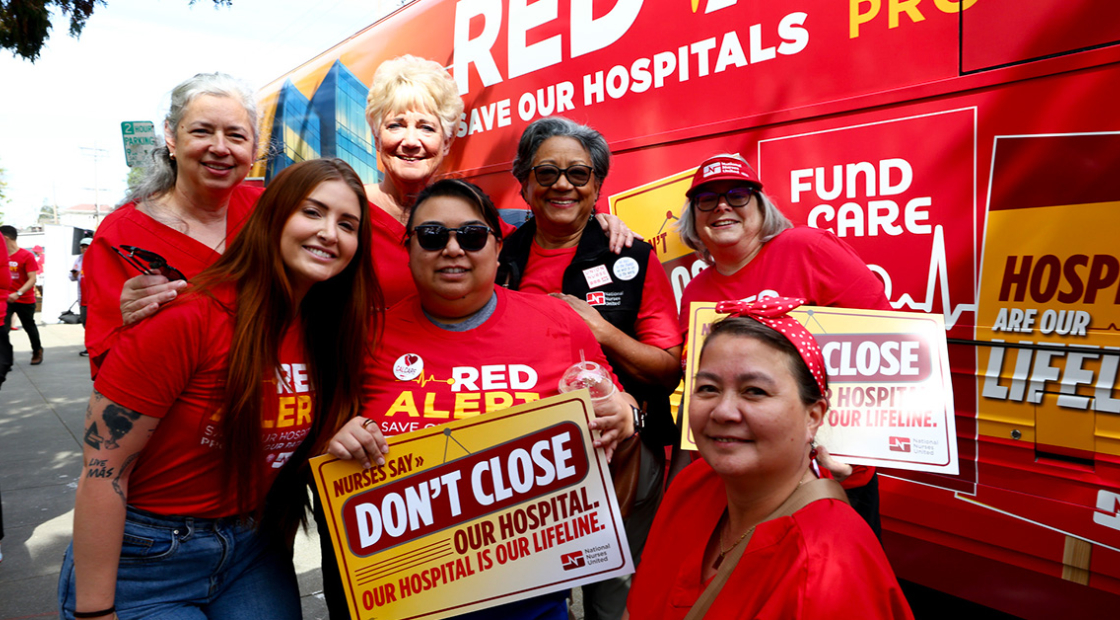 Group of nurses in front of Red Alert tour bus, smiling, holding sign "Don't close our hospital"
