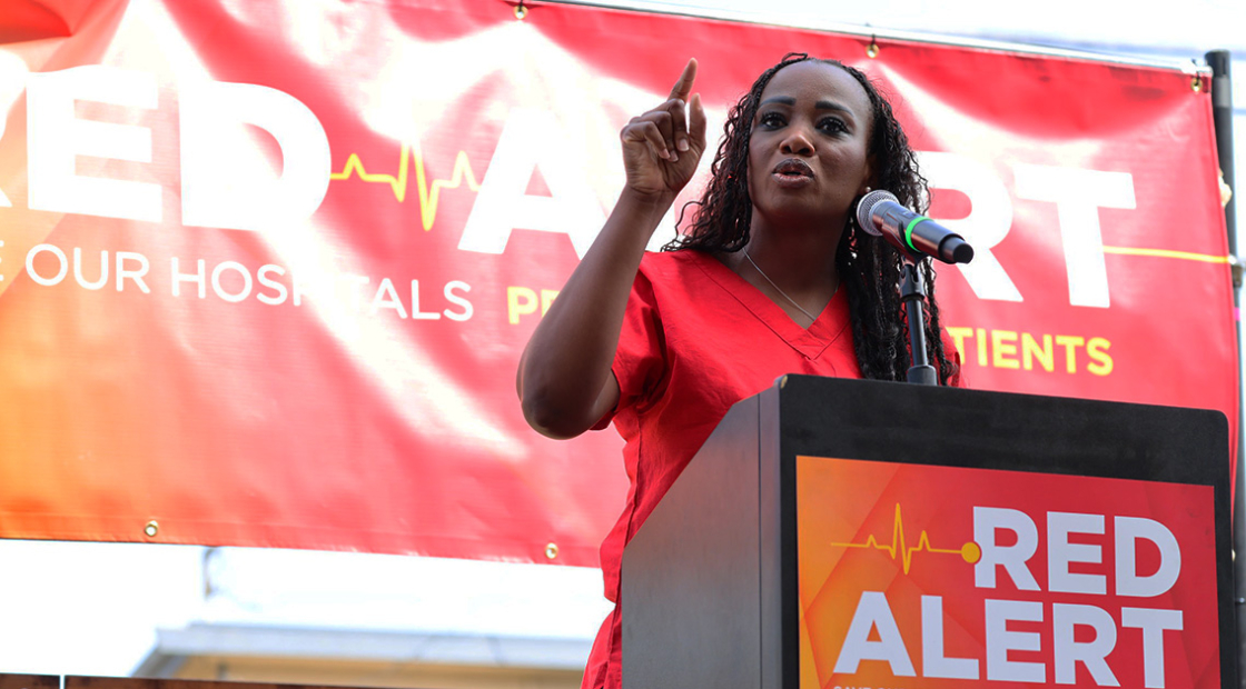 Nurse speaking at podium with banner behind "Red Alert: Save Our Hospitals Tour"