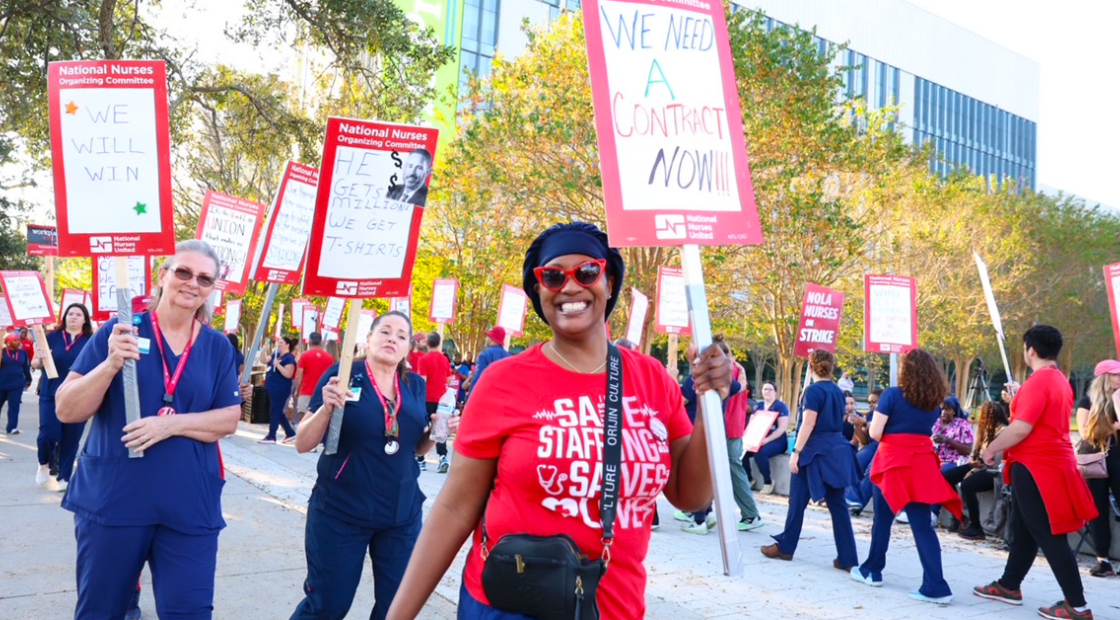 Nurses on picket line, smiling, one holds sign "We Need a Contract NOW!"