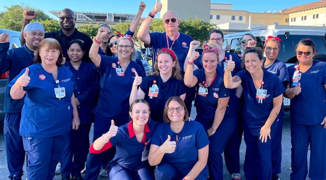 Large group of nurses outside hospital, wearing blud scrubs, smiling, and giving thumbs up