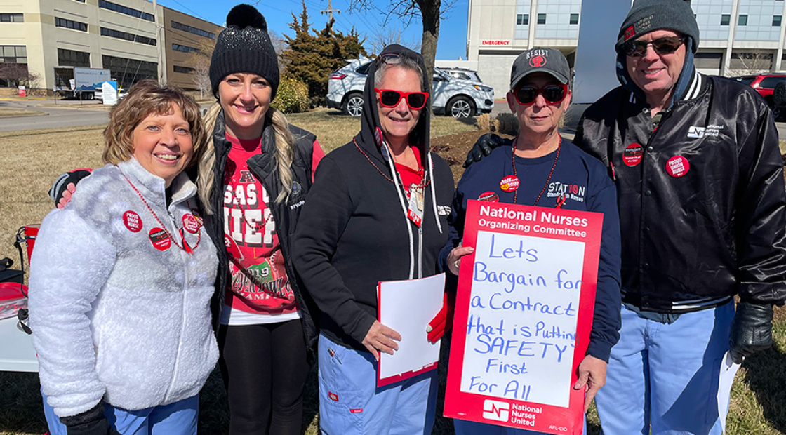 Five nurses outside hospital. One holds sign "Let's bargain for a contract that is putting SAFETY FIRST for all"