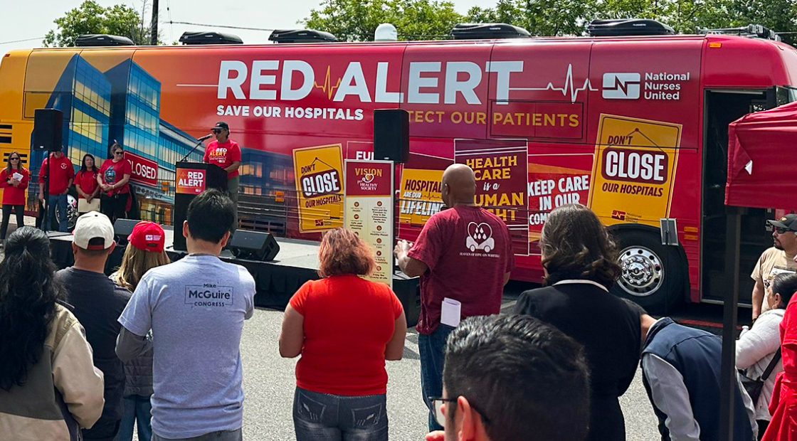 People speak at podium in front of Red Alert Tour bus, crowd watching