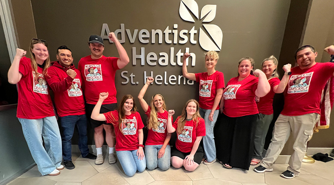 Group of nurses inside, in front of sign "Adventist Health", smiling, wearing red, and with raised fists