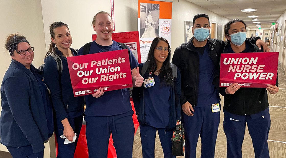Six nurses inside hospital, side-by-side, smiling, two holding signs "Union Nurse Power"