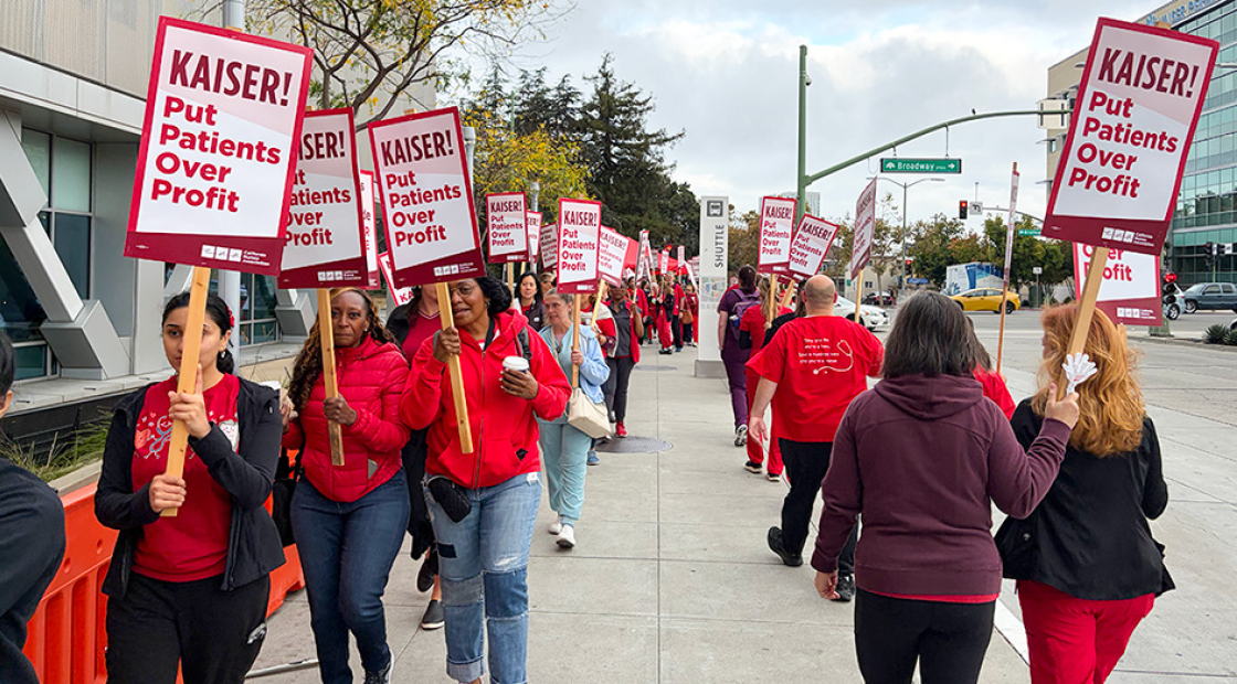 Nurses on picket line outside of hospital, holding signs "Kaiser: Put Patients Over Profits"