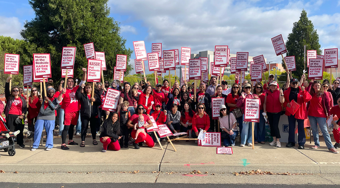 Large group of nurses outside holding signs "Kaiser: Put Patients Over Profit" and "Nurses at the Bedside, Kaiser at the Bank"